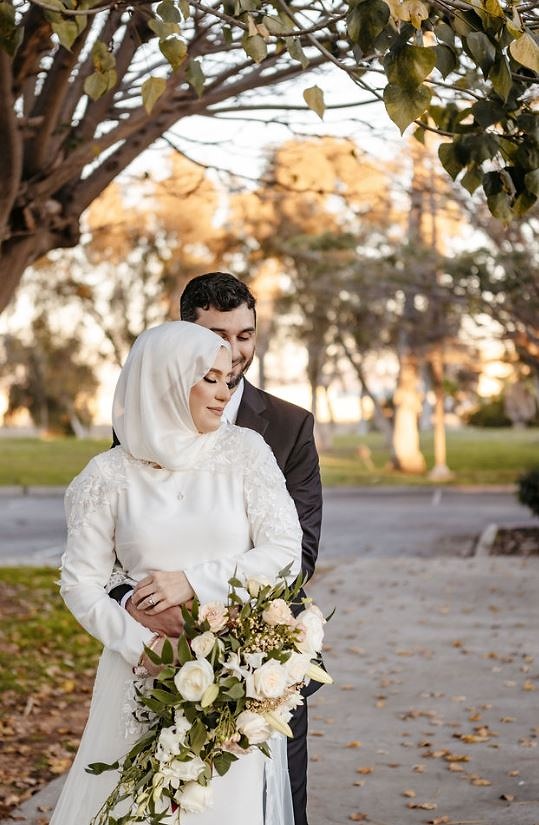 Bride in a hijab and long-sleeved gown held by her groom, holding a white rose bouquet