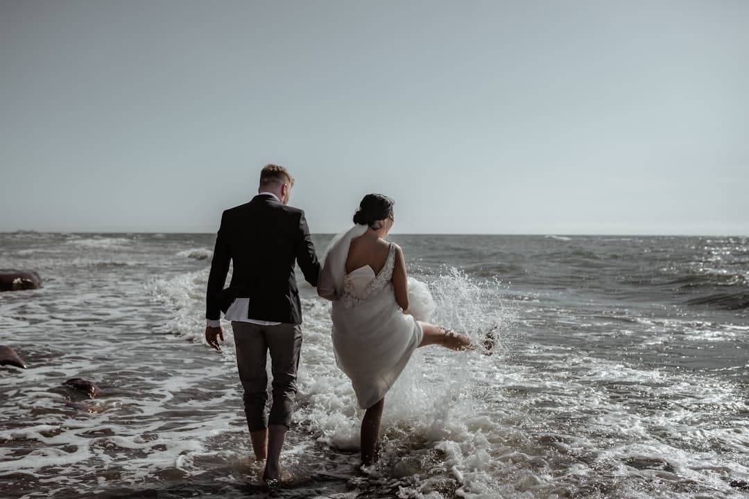 Bride and groom walking hand in hand into the surf, she kicks up a spray of water with her hem gathered in one hand