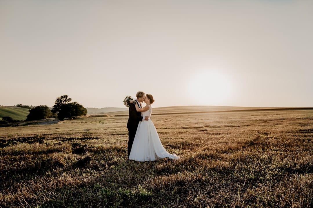 Bride and groom kissing alone in a wide harvested field, the low sun flaring across the stubble behind them