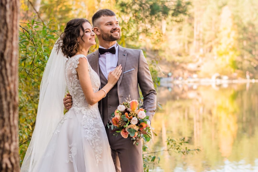 Bride and groom by a lake in autumn light, she leans into his shoulder while he holds a bouquet of peach and coral blooms