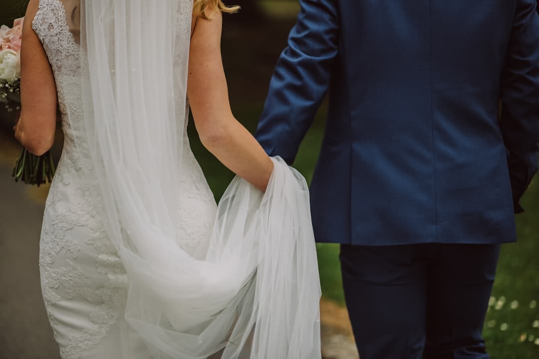 Detail from behind of a bride's lace gown and long veil gathered in her hand as she walks beside her groom
