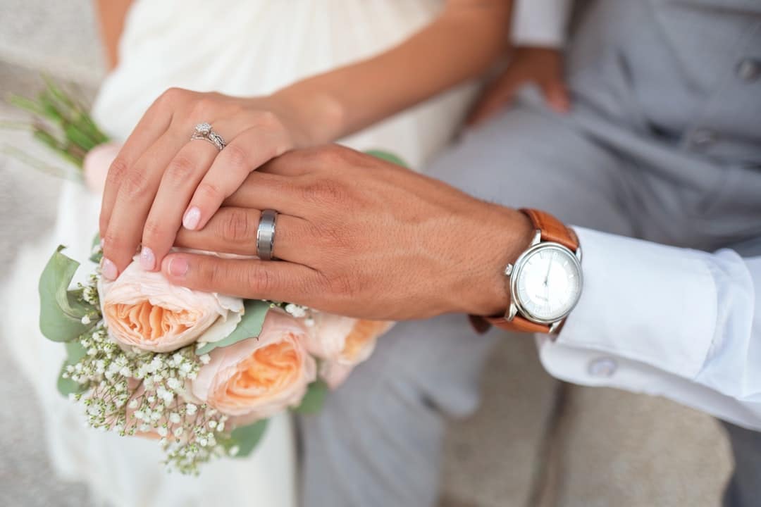 Couple's hands resting together on a bouquet of peach garden roses, her engagement ring and his matte band in close view