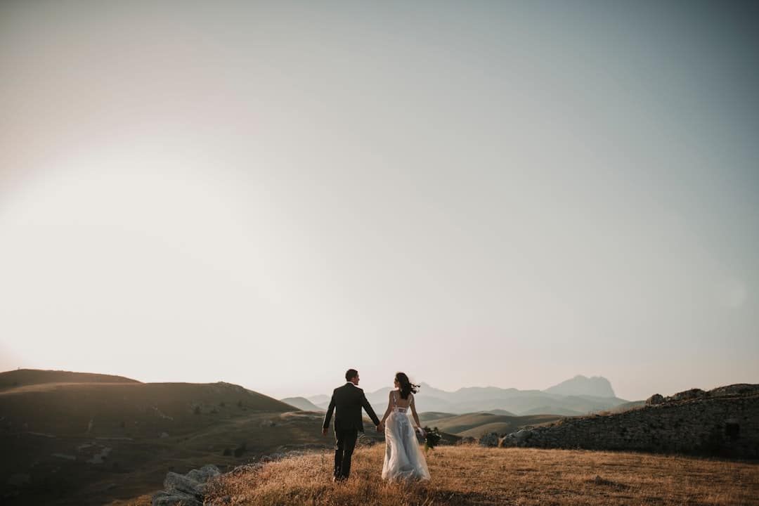 Bride and groom walking hand in hand along a high ridge at sunset, mountains fading into haze ahead of them