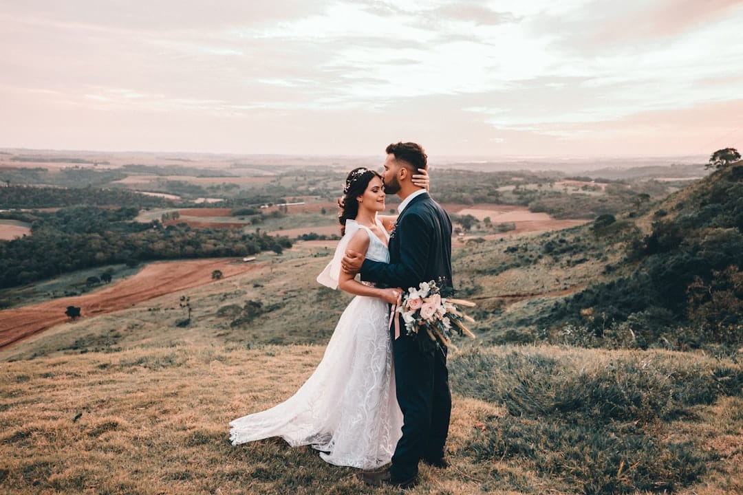 Bride and groom in a quiet embrace on a hilltop at sunset, her bouquet of pale roses between them and farmland beyond