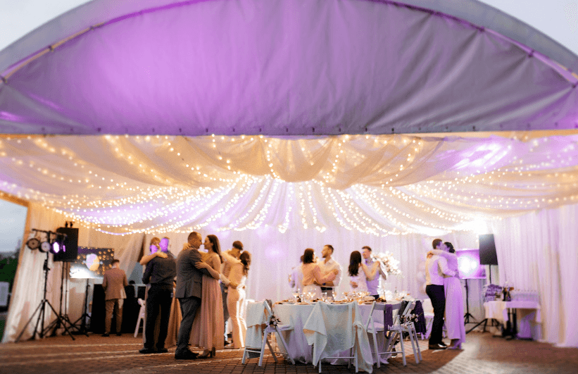 Guests slow-dancing under a tent draped with fairy lights and lit purple at an evening reception