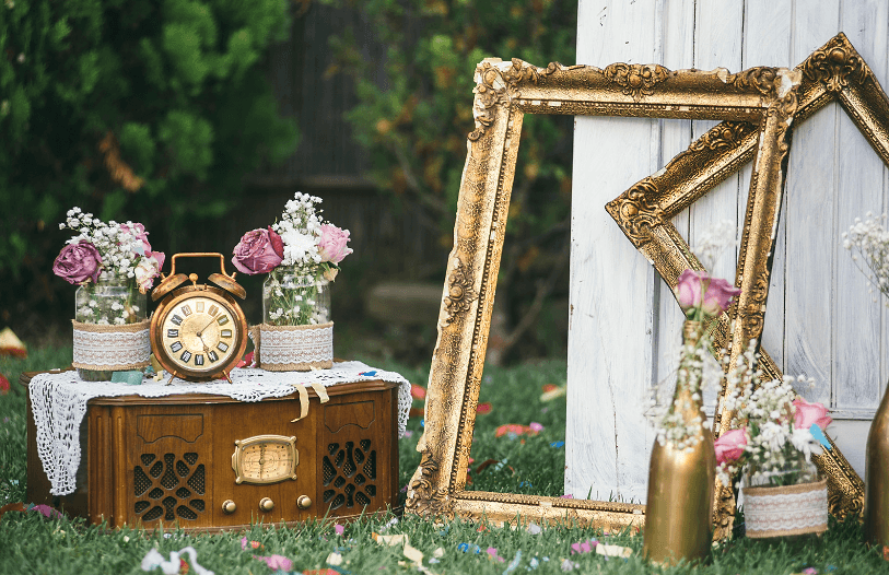 Vintage wedding decor on a lawn: gilded empty frames, a copper alarm clock on an old radio, and roses in lace-wrapped jars