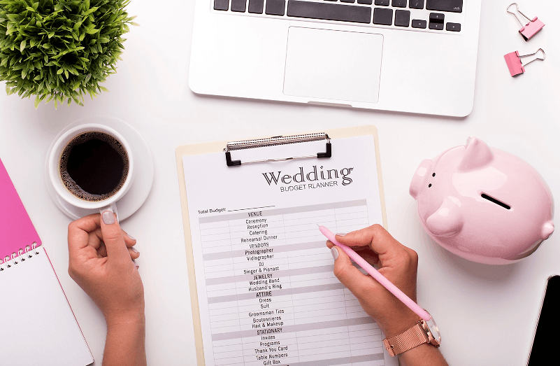 Hands filling in a wedding budget planner on a clipboard beside a cup of coffee and a pink piggy bank