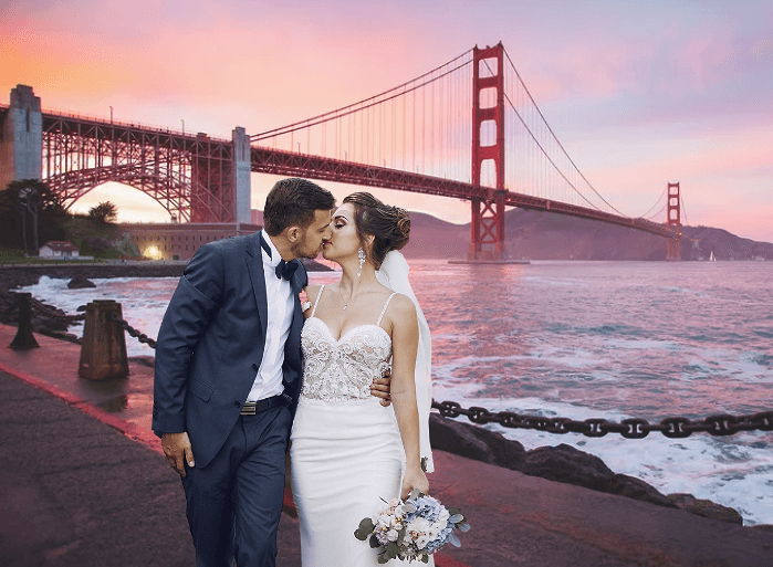 A bride and groom kiss on the waterfront beneath the Golden Gate Bridge at a pink sunset