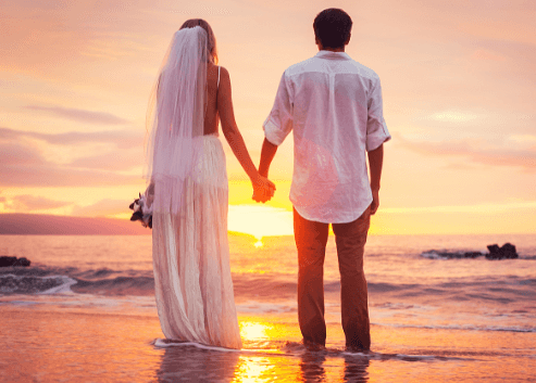 Bride and groom standing hand in hand at the water's edge, watching the sun set over the ocean in front of them