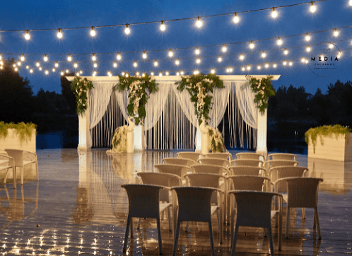 Evening ceremony set for a small guest list under canopies of string lights, a draped arch dressed with greenery at the water's edge