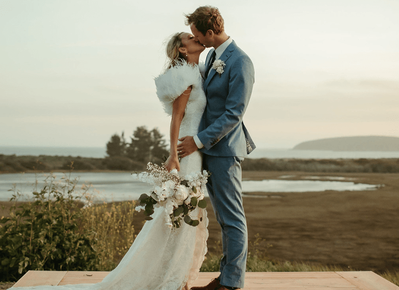 A couple kiss on a wooden deck above the Bodega Bay wetlands at dusk, the bride holding white roses