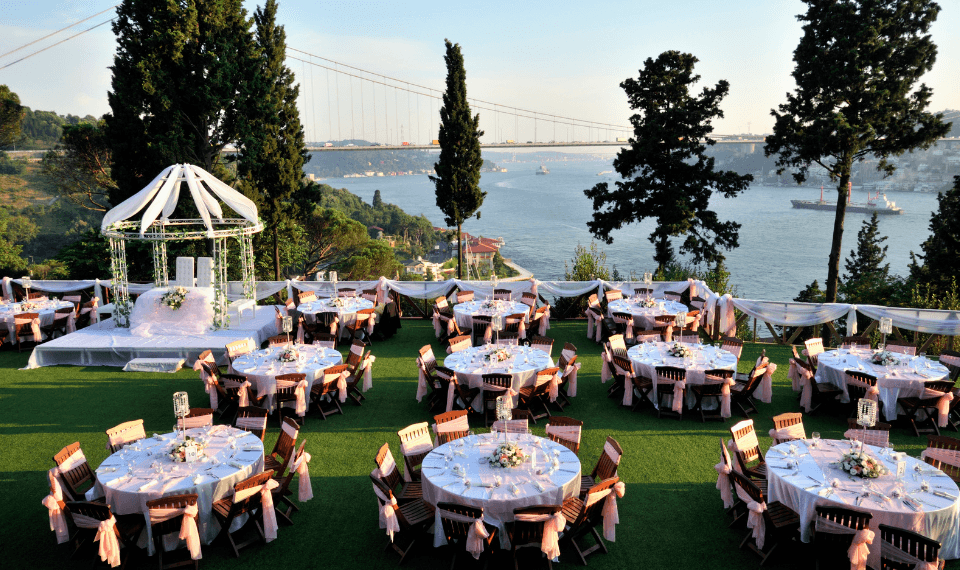 Outdoor reception of round tables dressed in white and blush on a lawn above the water, a draped ceremony pavilion at one end
