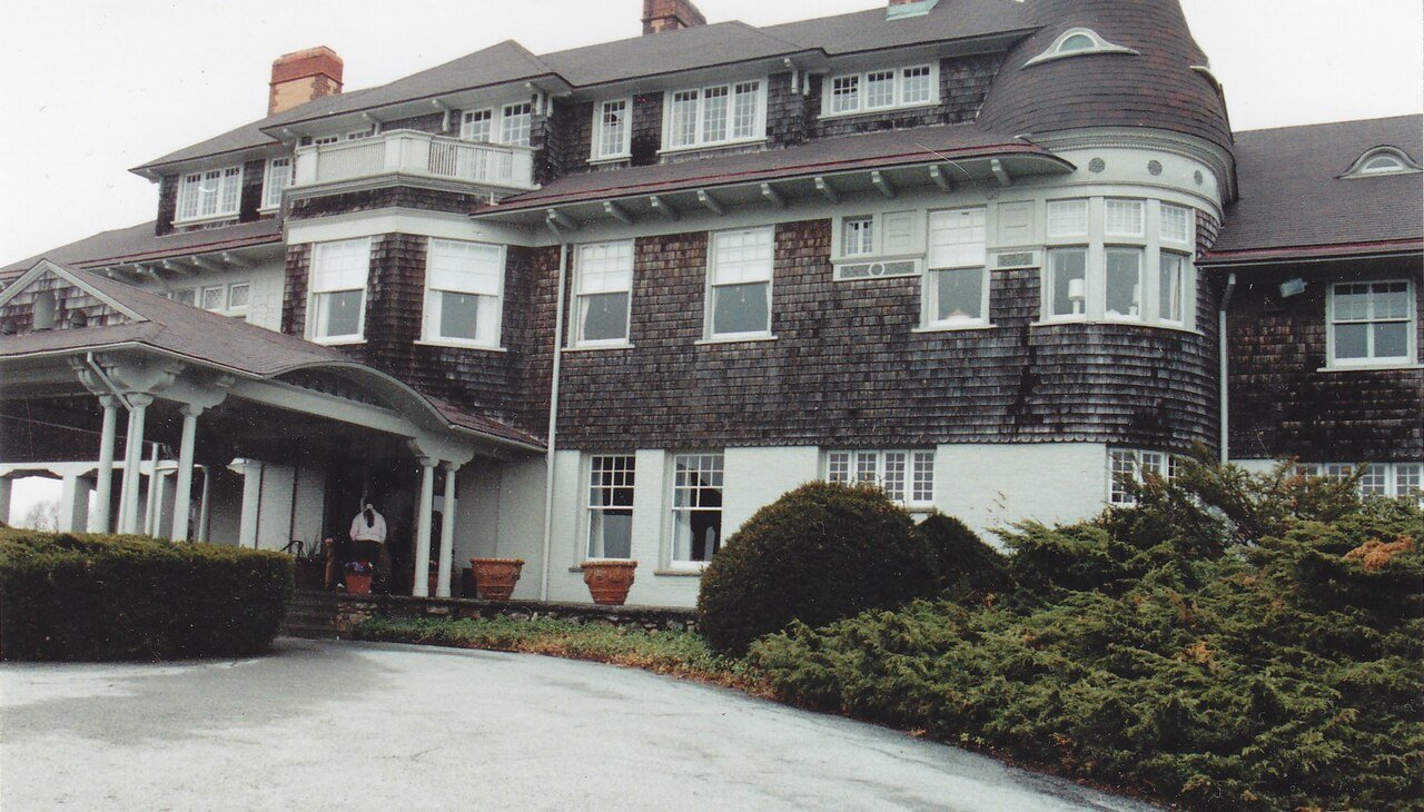 Grand shingle-style mansion with a covered porch, a curved turret and clipped hedges along the drive