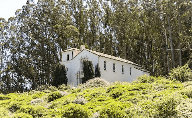 The white Spanish-style Presidio Chapel on a green hillside beneath tall eucalyptus trees