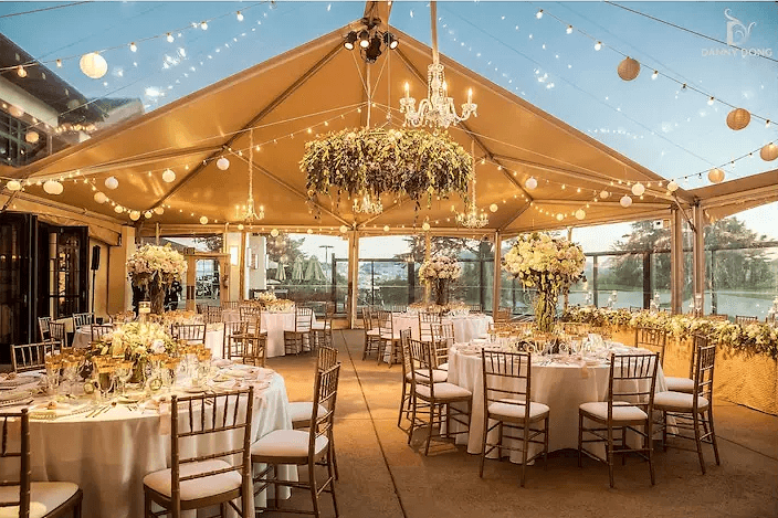 Reception laid out under a clear tent at the Presidio Log Cabin, with a chandelier, a hanging greenery wreath and string lights at dusk