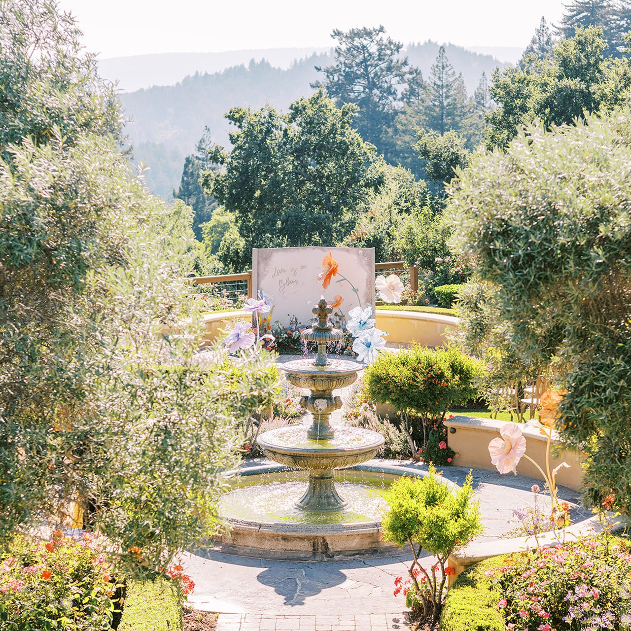 Tiered stone fountain in the garden at Regale Winery & Vineyards, with a floral Love is in Bloom backdrop and forested hills behind
