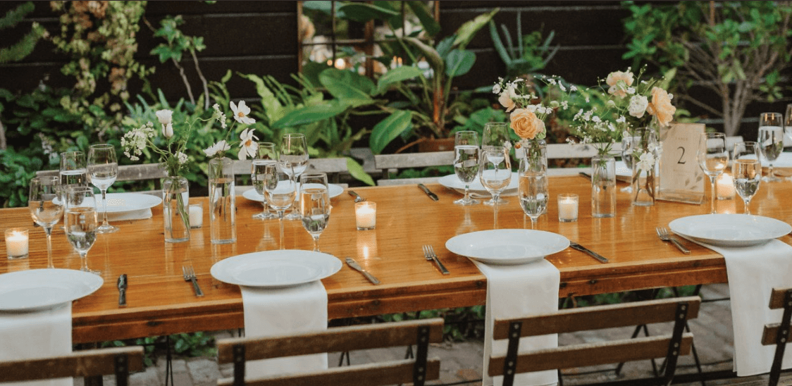 Long wooden table set for dinner in the leafy garden courtyard at Stable Café, with white roses, candles and folded linen napkins