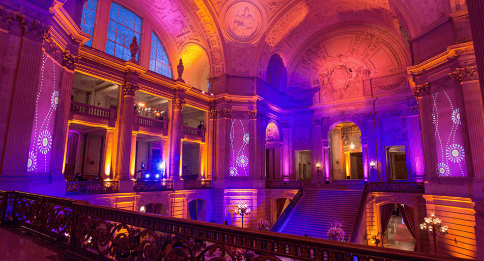 The rotunda of San Francisco City Hall lit purple and orange for an evening event, seen from an upper balcony