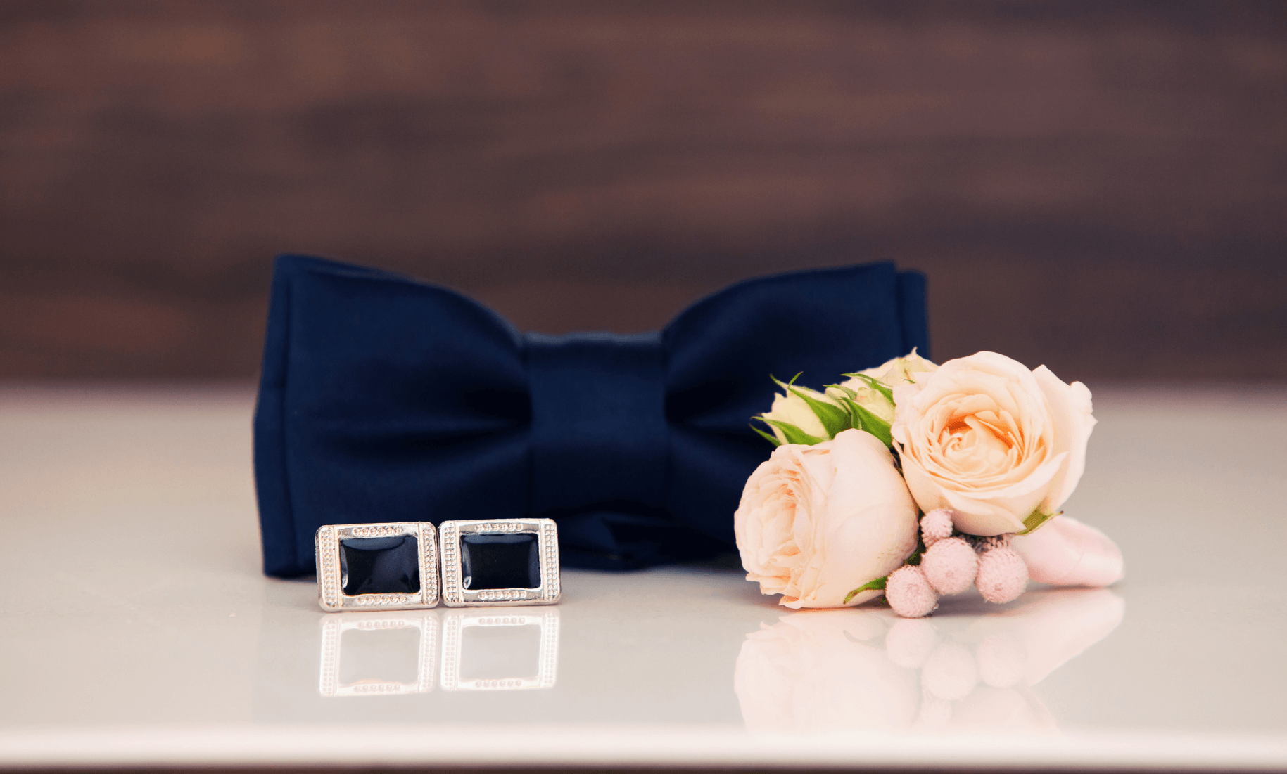 Navy bow tie, a pair of square silver cufflinks and a peach rose boutonniere on a white surface