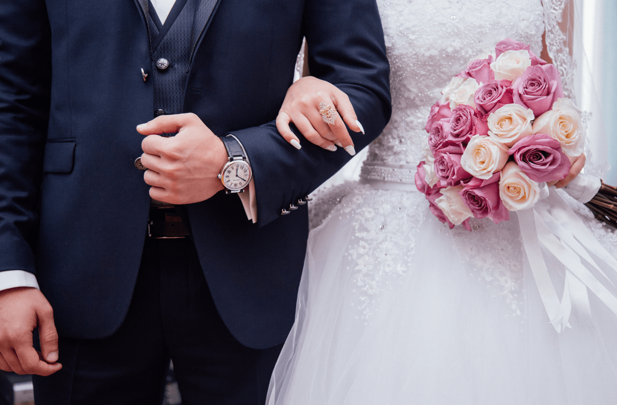 Bride holding the arm of a groom in a navy three-piece suit, with a bouquet of pink and white roses