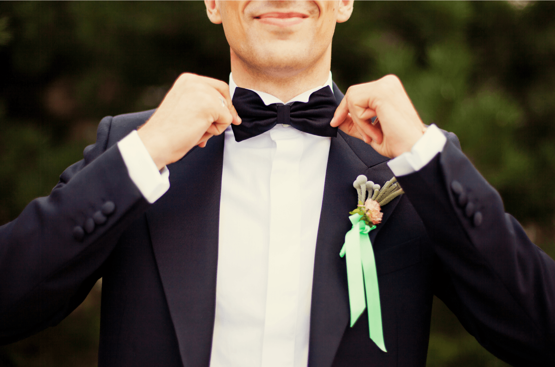 Groom straightening a black bow tie over a white shirt, a boutonniere with mint-green ribbon on the lapel