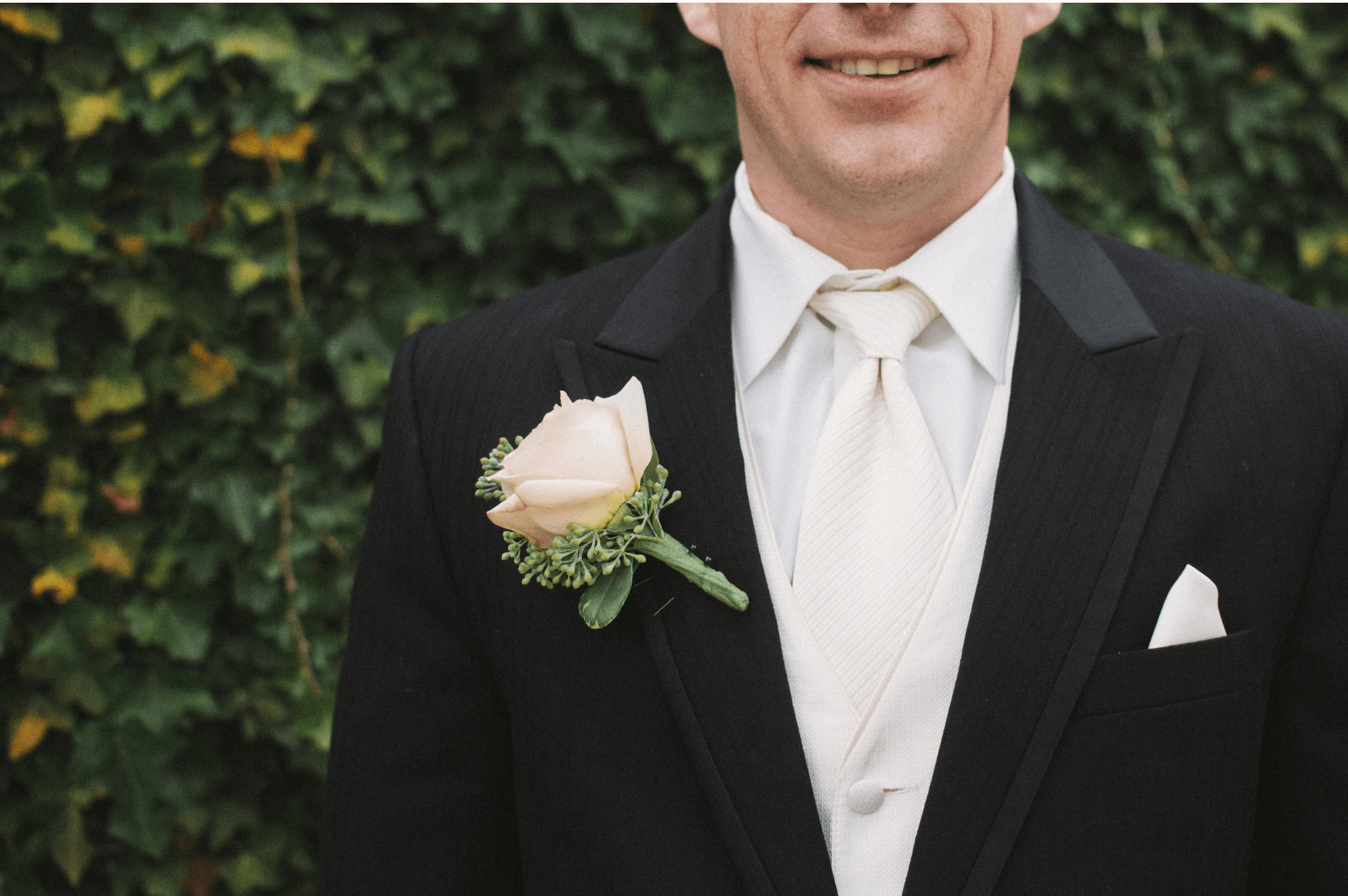 Close-up of a groom in a black tailcoat with an ivory cravat, waistcoat and pocket square, a cream rose boutonniere on his lapel