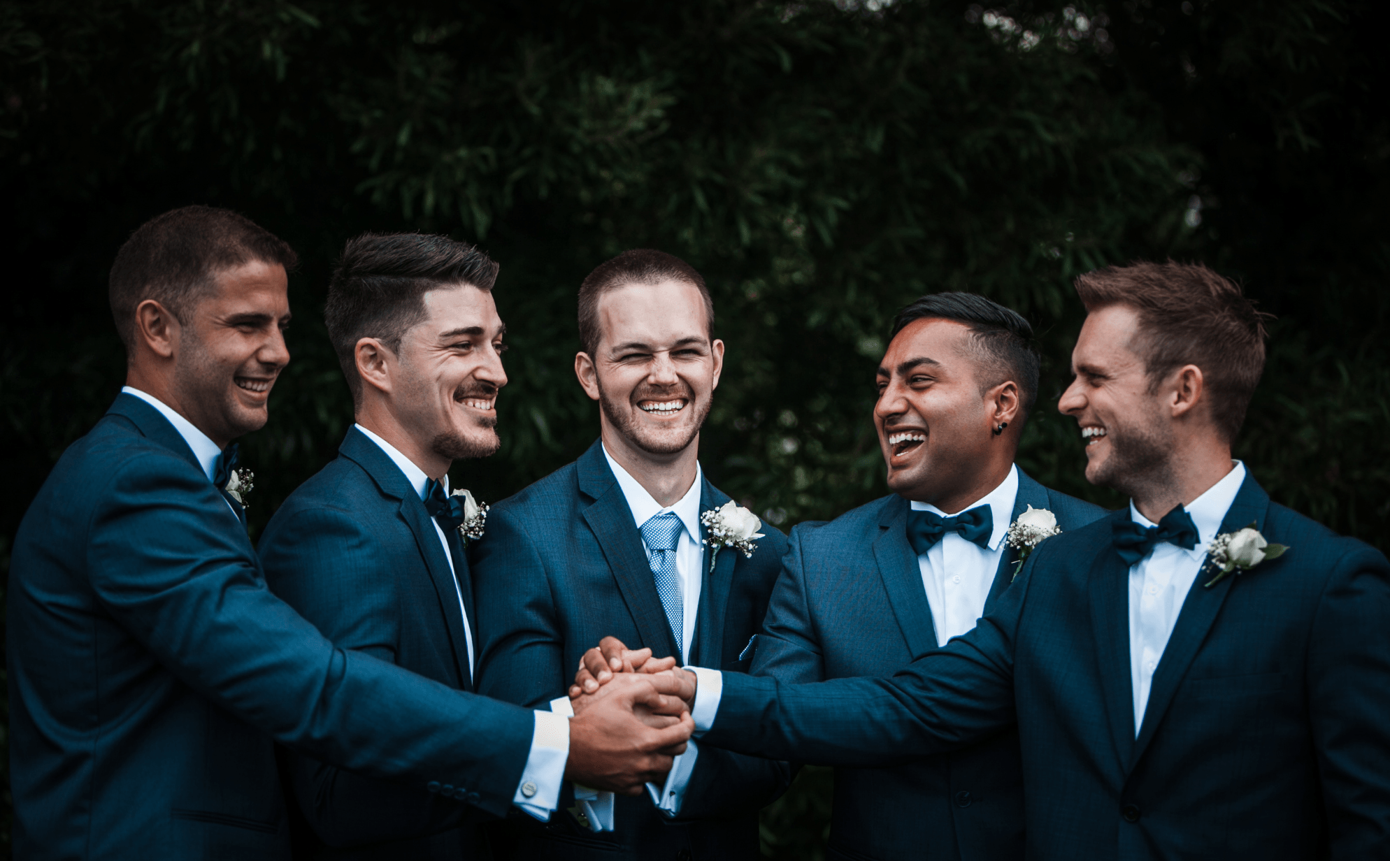 Groom and four groomsmen in navy suits with white rose boutonnieres, laughing together