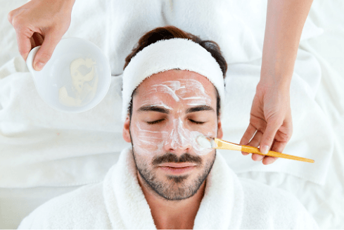 Man lying back in a towelling robe and headband while a facial mask is applied with a brush