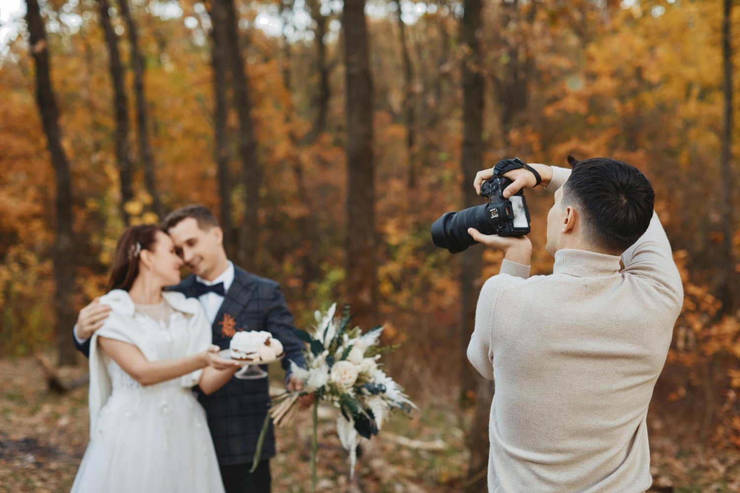 Photographer raising his camera to a bride and groom holding a small cake among autumn trees