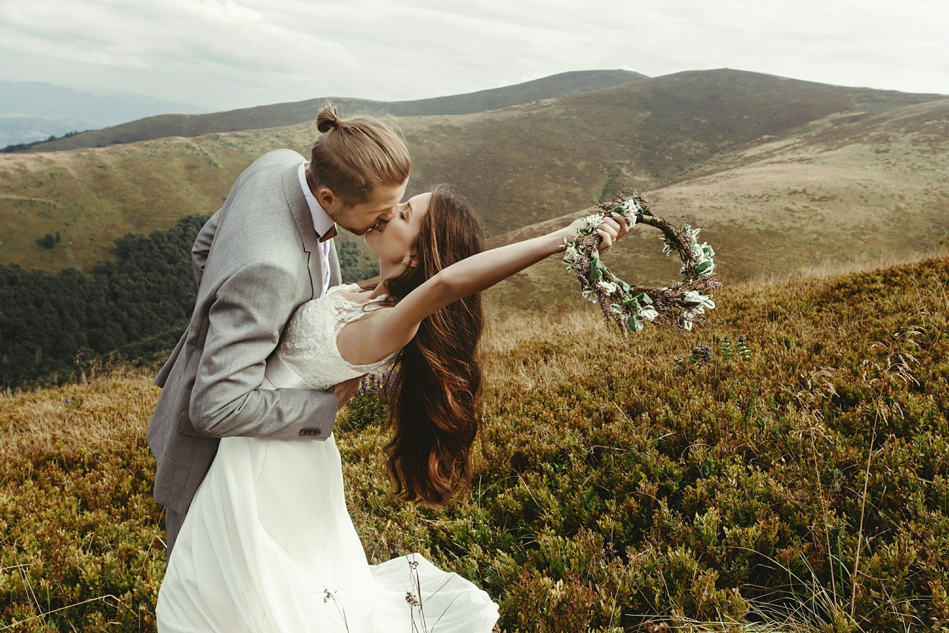 Groom dipping his bride for a kiss on an open mountain ridge, she holds a flower crown out at arm's length