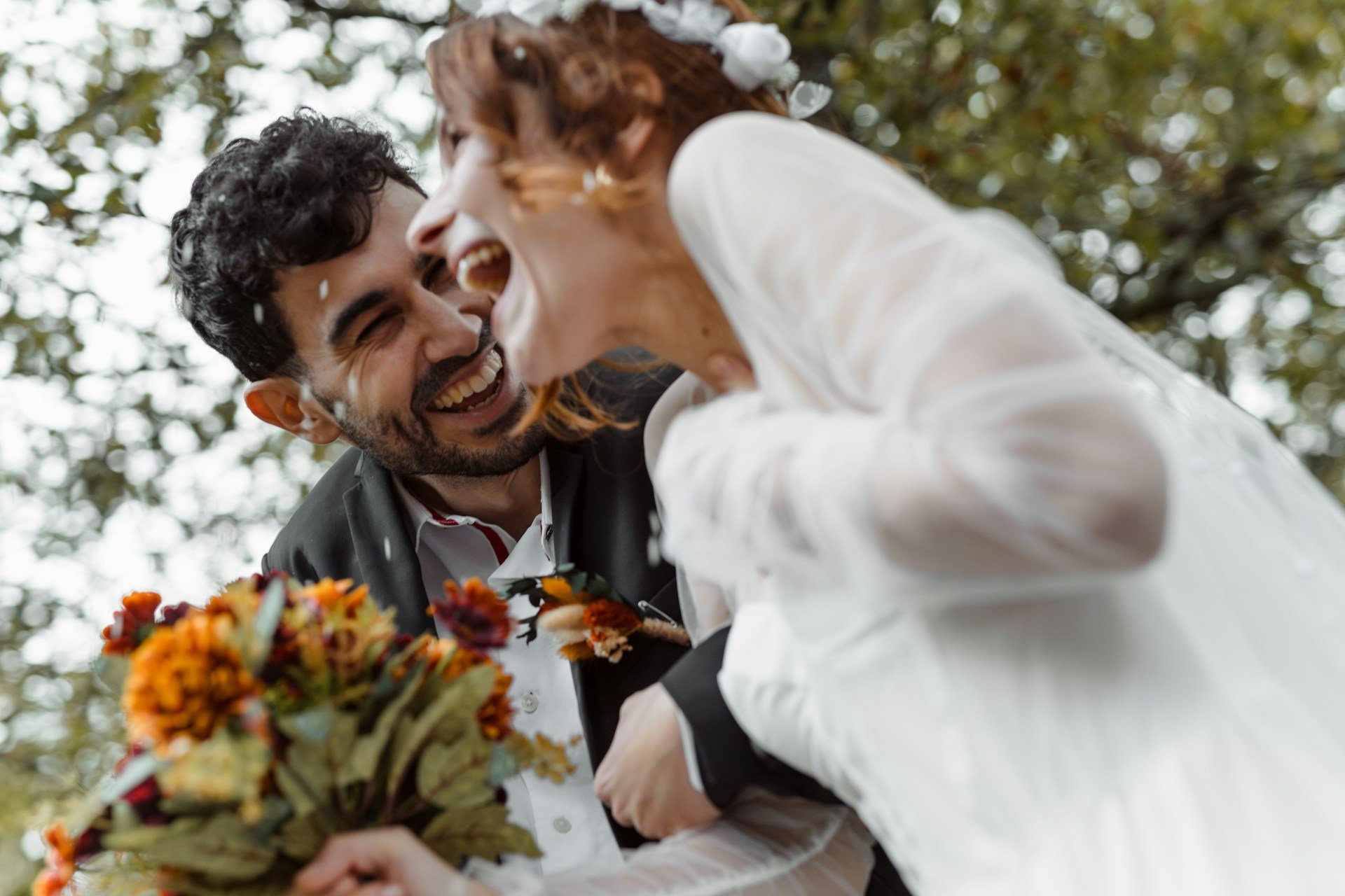 Bride and groom caught mid-laugh under falling confetti, her autumn bouquet of orange and burgundy blooms held in the foreground