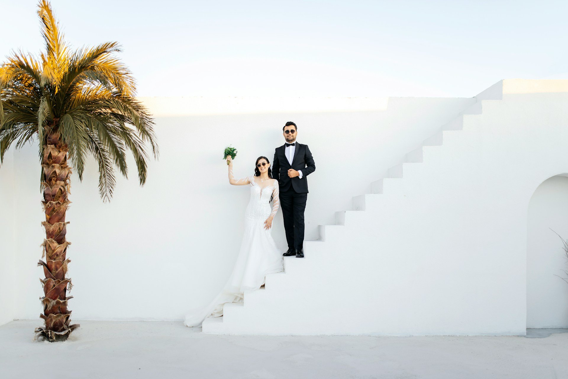 Bride and groom in sunglasses posed on whitewashed steps beside a palm tree, she raises her bouquet overhead