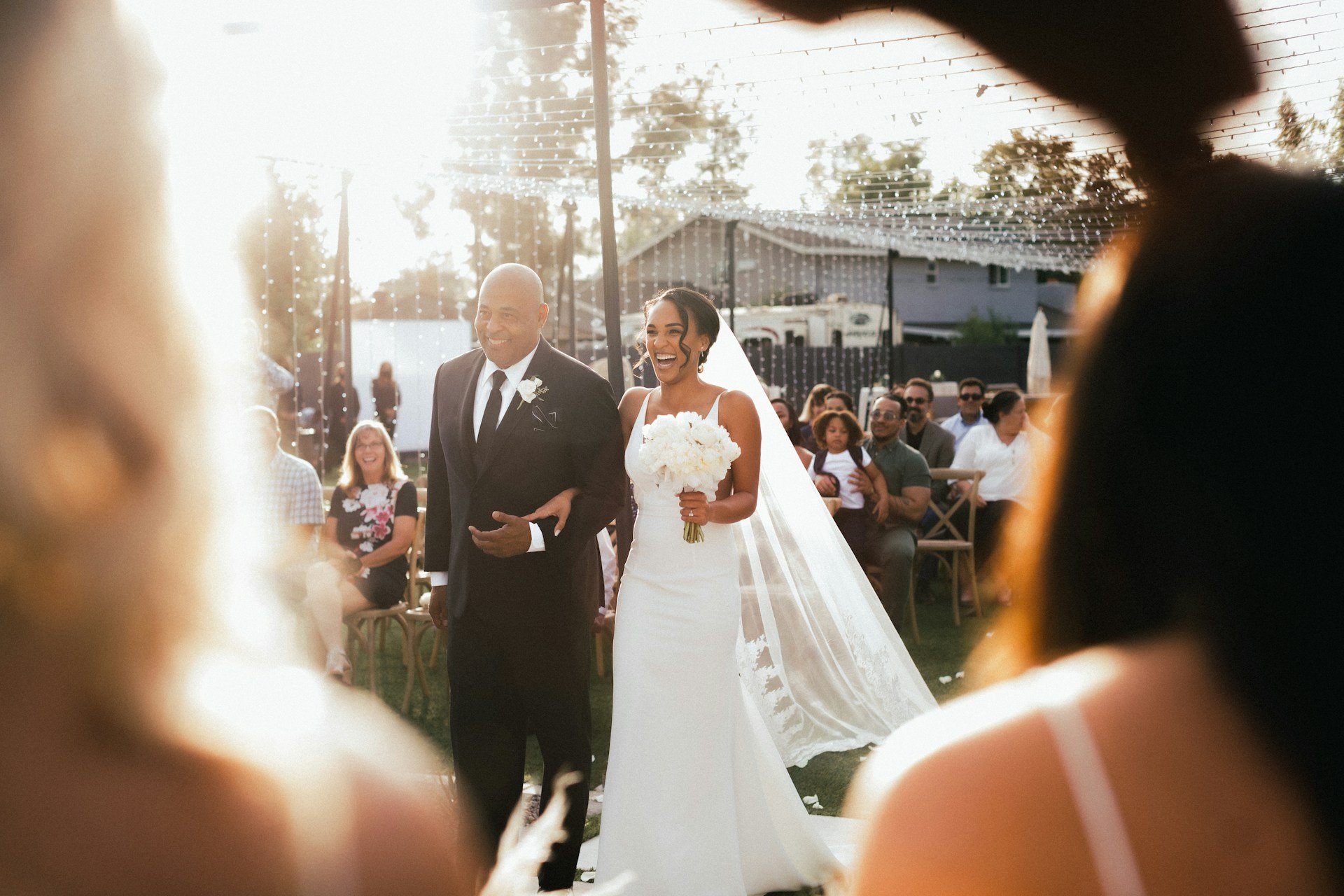 Bride laughing as she walks down an outdoor aisle on her father's arm, guests seated beneath canopies of fairy lights