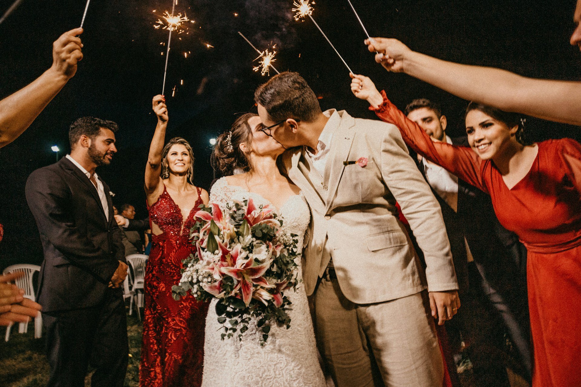 Newlyweds kissing under an arch of sparklers held by guests at night, her lily bouquet held between them