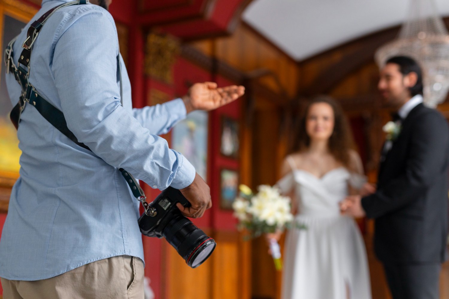 Wedding photographer gesturing to direct a couple during their ceremony, camera in hand and the pair softly out of focus beyond him