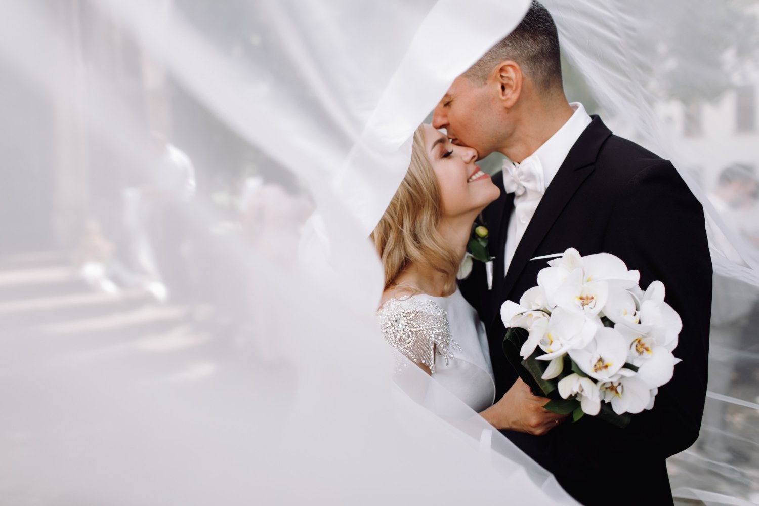 Bride and groom sheltered under her veil as it lifts in the wind, she holds a bouquet of white orchids against his tuxedo