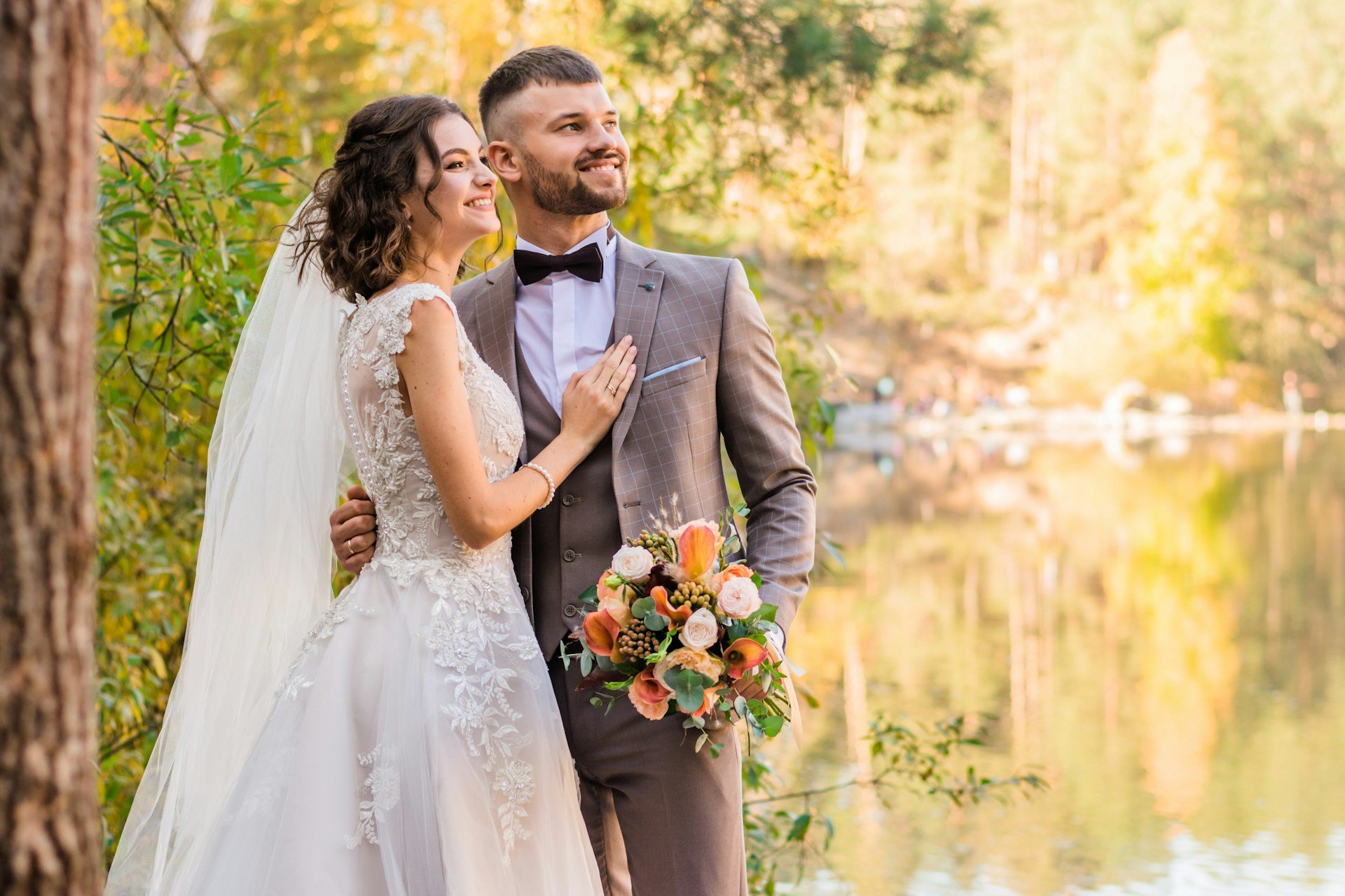 Bride and groom by an autumn lake, she rests a hand on his checked brown suit and he holds a bouquet of peach roses and calla lilies