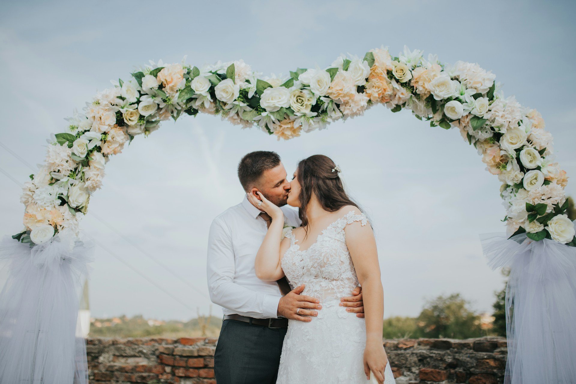 Bride and groom kissing beneath a tall arch of cream and peach roses, open countryside behind them