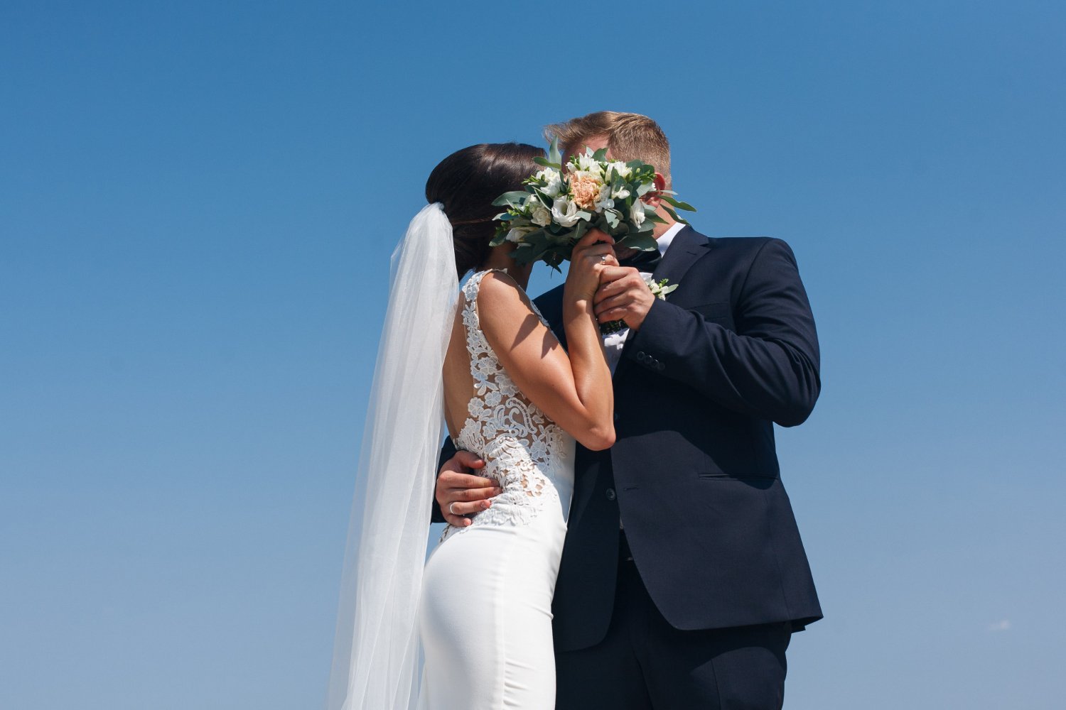 Bride and groom kissing behind her raised bouquet against a clear blue sky, her veil falling the length of her back