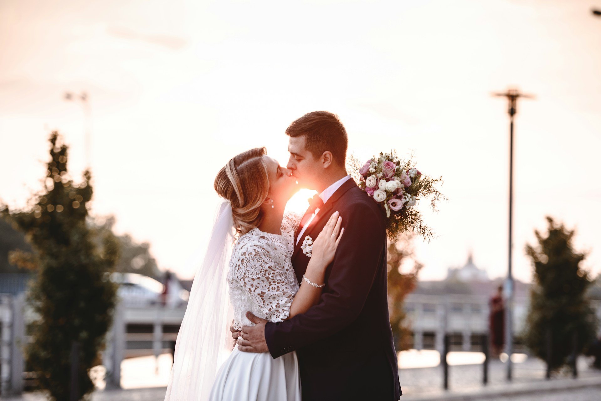 Bride and groom kissing at sunset with the light flaring between them, her lace sleeves and long veil catching the glow