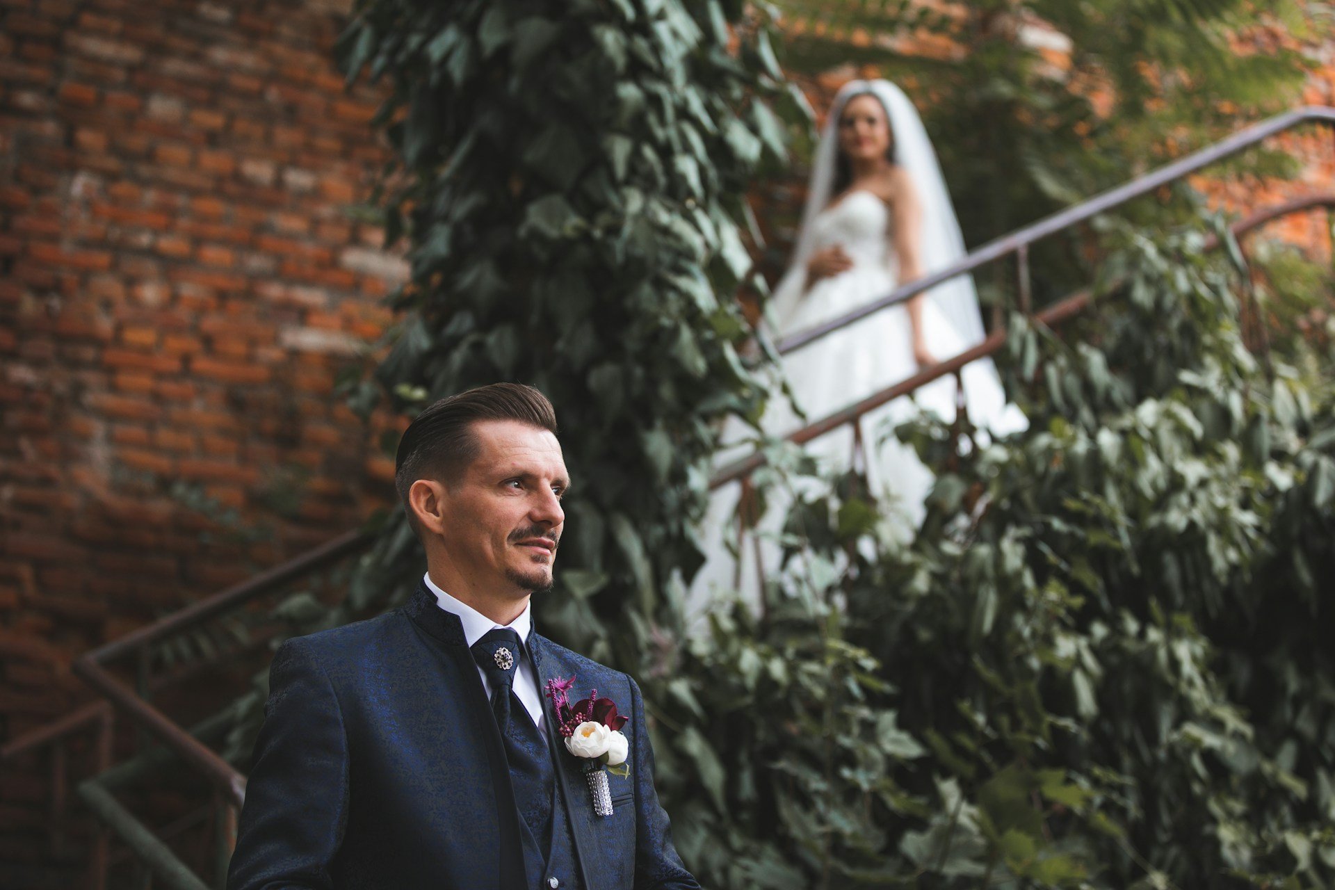 Groom in a patterned navy suit waiting in the foreground while his bride watches from an ivy-covered staircase behind him