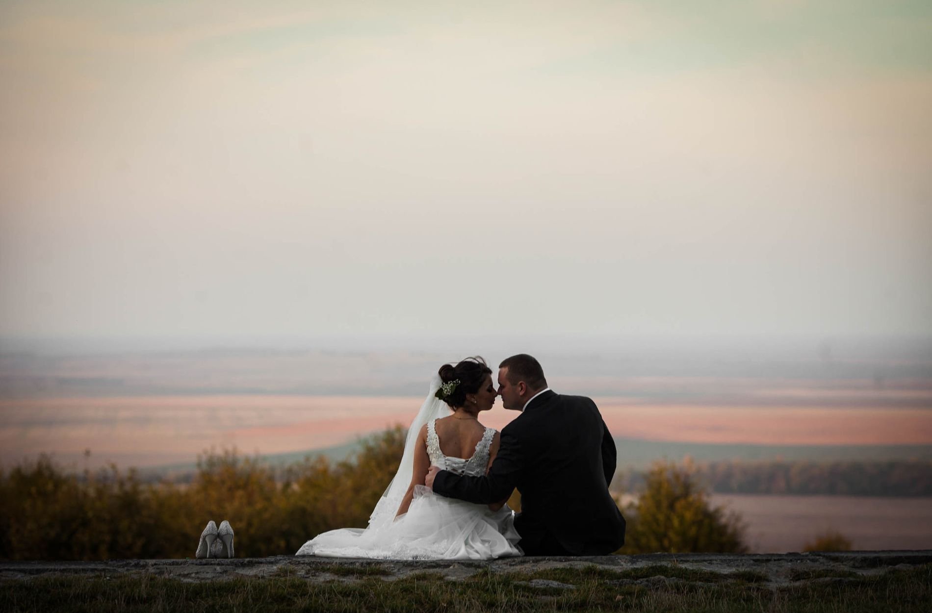 Bride and groom sitting together on a clifftop at dusk, foreheads touching, her veil trailing across the rock with open countryside behind them