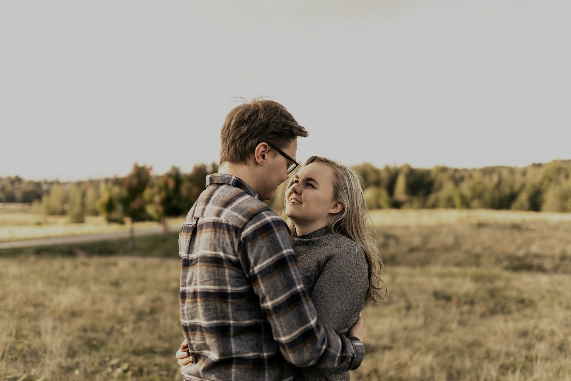 Engaged couple holding each other in an open field at golden hour, she looks up at him mid-laugh