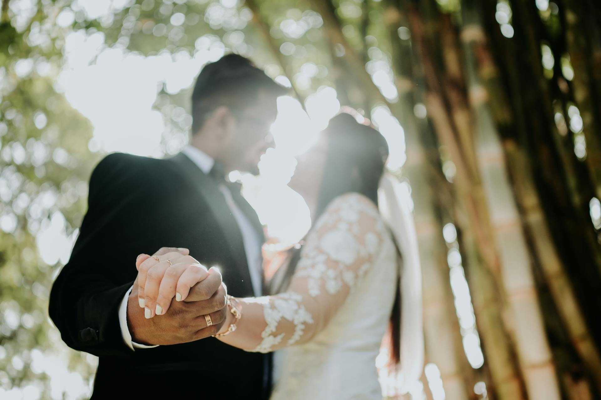 Backlit candid of a bride and groom leaning in to kiss, their clasped hands in the foreground showing her engagement ring and his wedding band