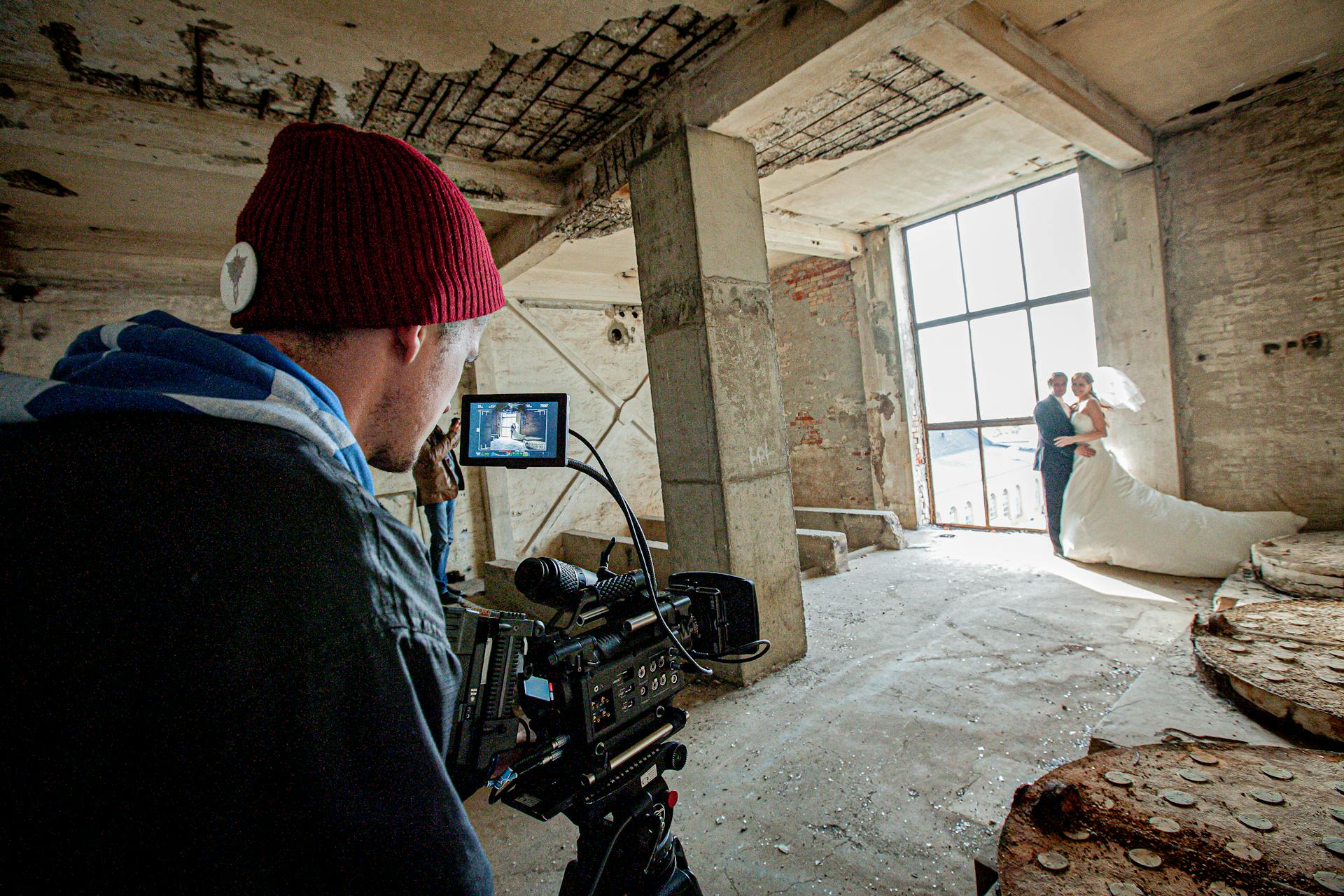 Wedding videographer in a red beanie filming a bride and groom framed in a tall window of a derelict concrete building, the shot visible on his monitor