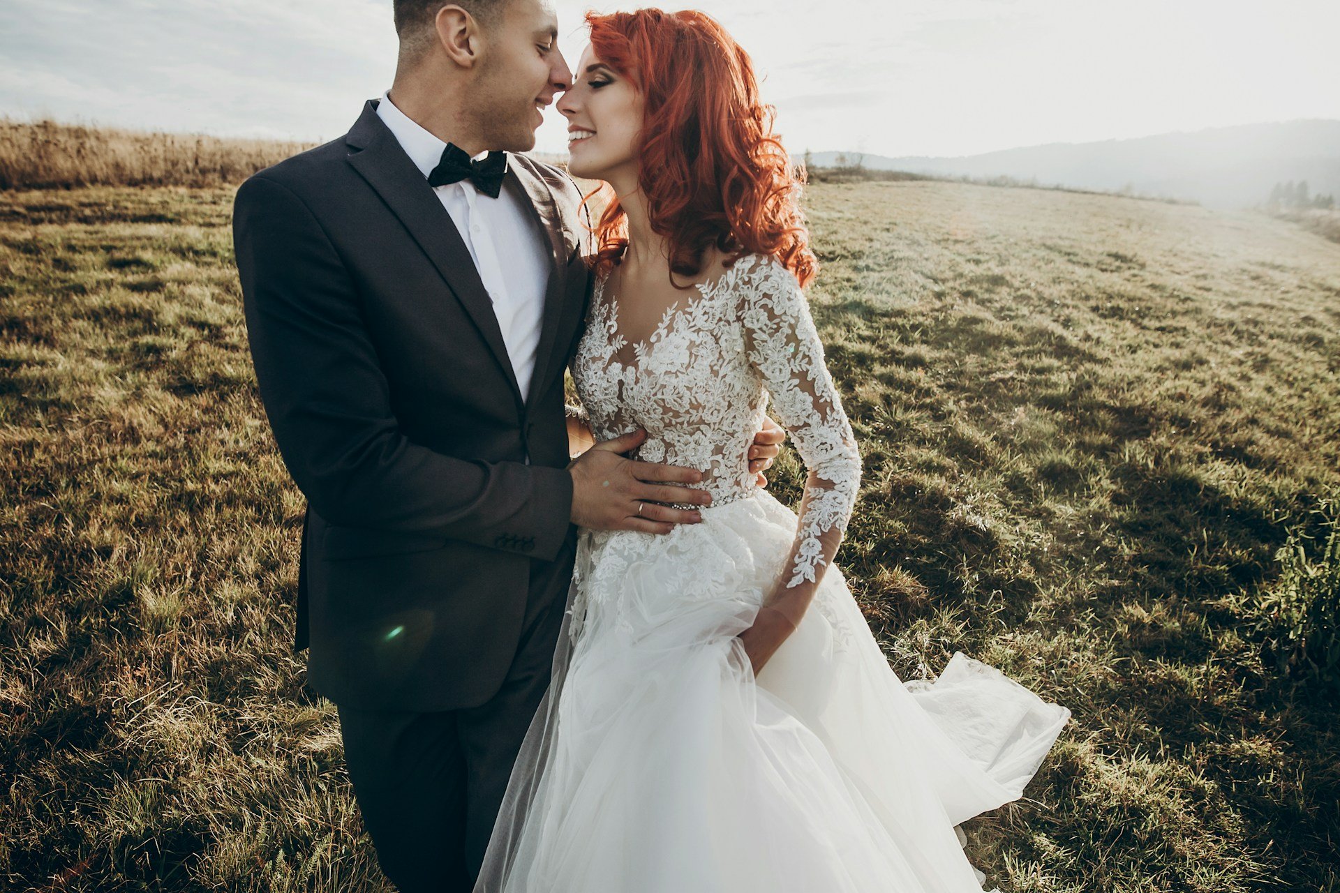 Red-haired bride in a long-sleeved lace gown leaning into her groom in a sunlit hillside meadow, both smiling with their eyes closed