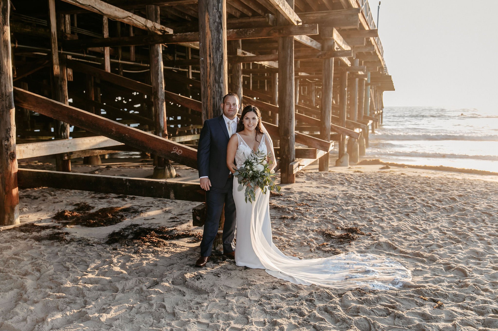 Bride and groom posing beneath a coastal pier at sunset, showing how to select a wedding photographer who can create timeless portraits in scenic beach locations.