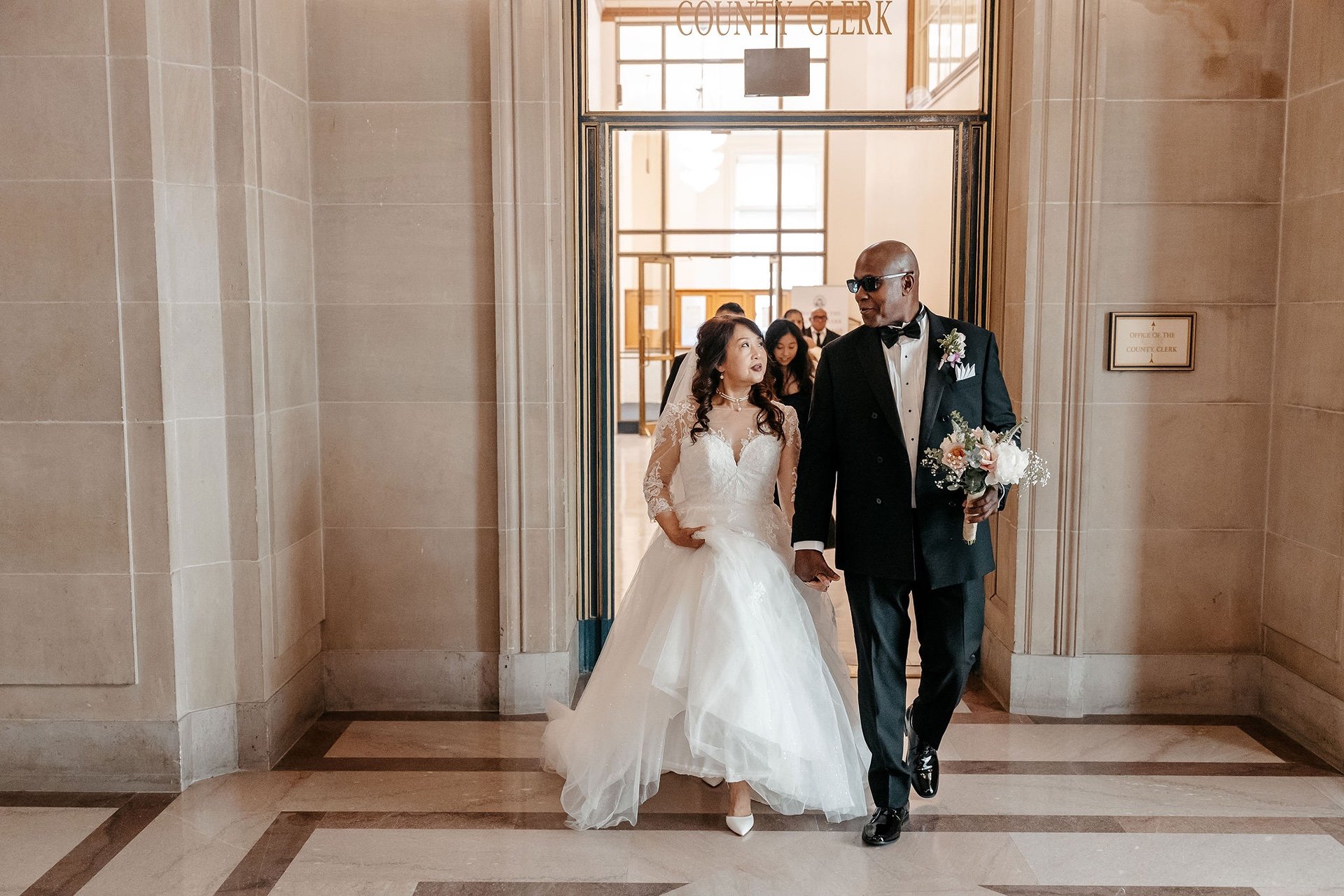 A bride and groom hold hands as they walk out of the County Clerk's office at San Francisco City Hall, the groom carrying her bouquet