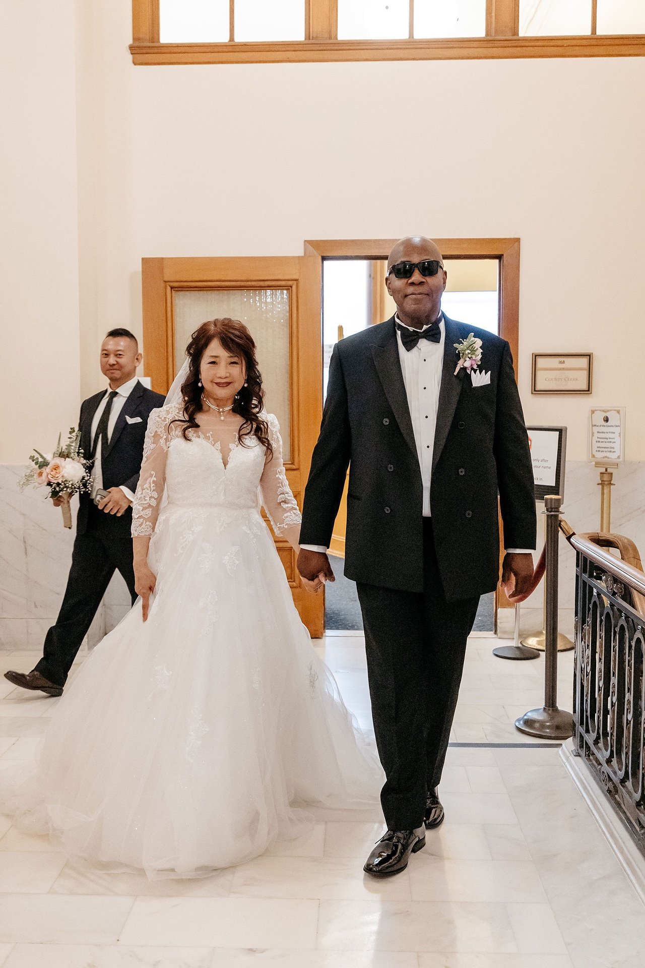 A bride and groom walk hand in hand past the County Clerk's office at San Francisco City Hall