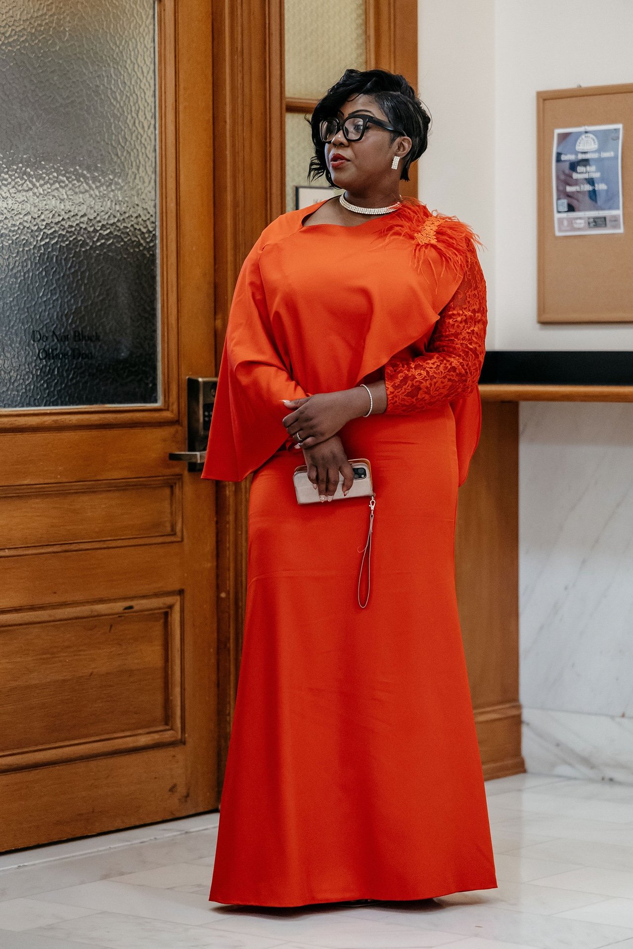 A wedding guest in a long red gown with a feathered shoulder and black glasses waits beside a wooden office door